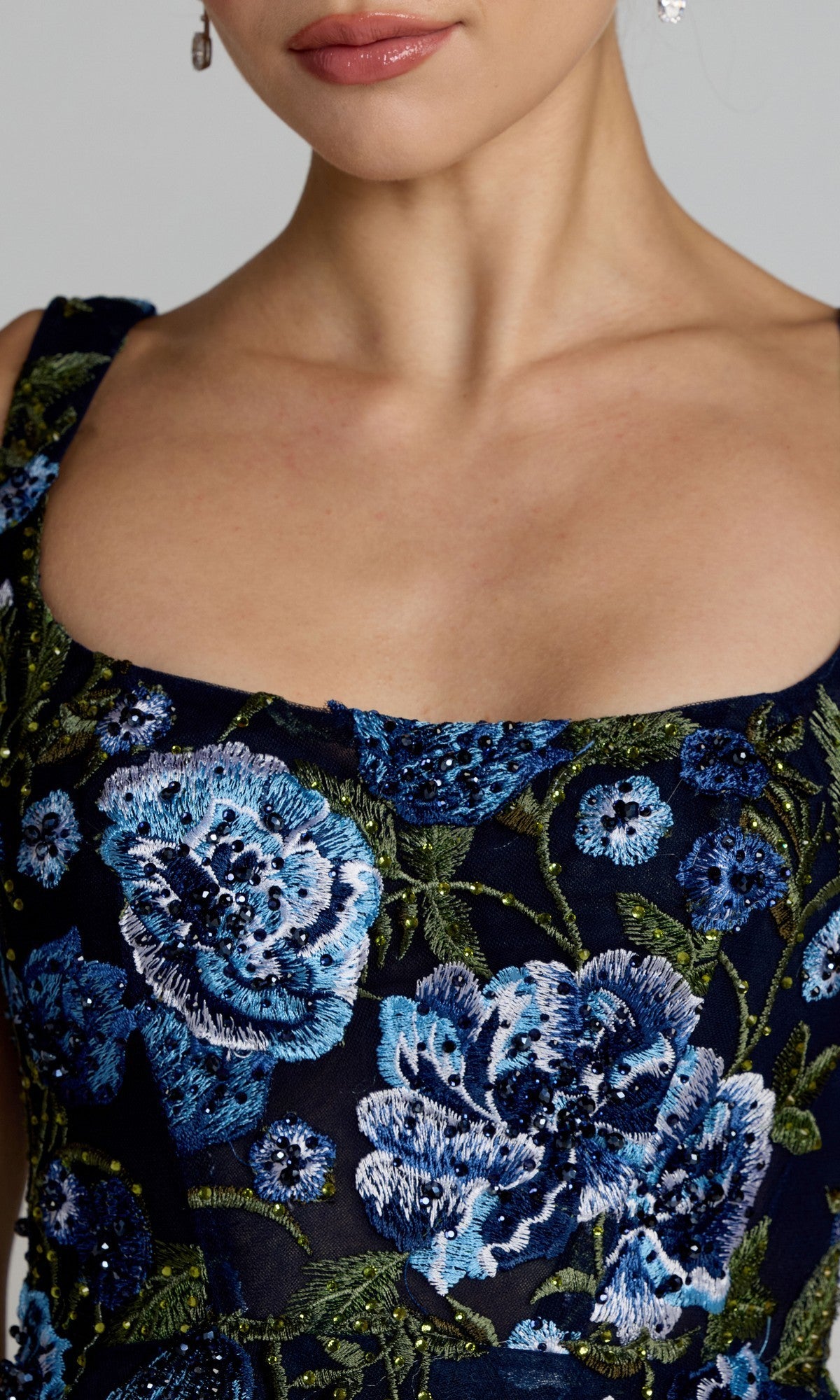 Close-up of a person wearing the Tea-Length Party Dress: Mac Duggal 21295, a sleeveless navy midi dress adorned with blue floral embroidery and beaded details on dark fabric. The person also wears small dangling earrings.